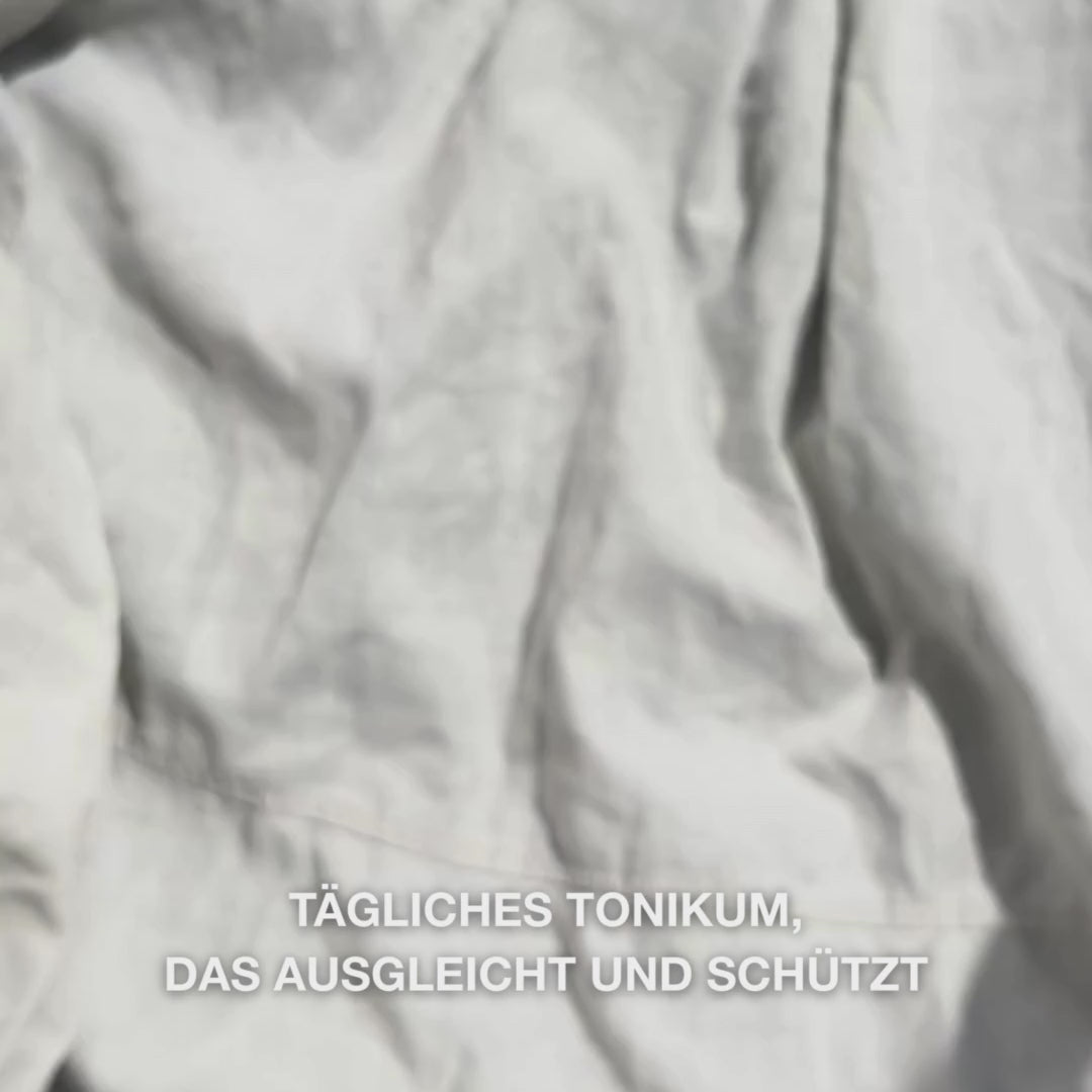 Nahaufnahme einer Hand, die einige Tropfen des klaren, wässrigen Face Tonic mit Teebaumöl auf ein Wattepad gibt und es dann sanft über die gereinigte Gesichtshaut streicht: die leichte, erfrischende Flüssigkeit beruhigt, reinigt tief, spendet Feuchtigkeit und hinterlässt die Haut erfrischt und optimal vorbereitet für eine Creme.